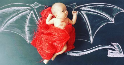 Directly above shot of baby girl lying amidst devil wings drawn on blackboard