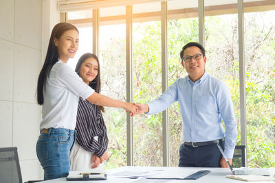 Smiling business people shaking hands while standing by colleague and table