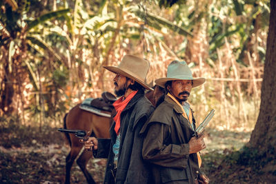 Men holding gun wearing hat standing with horse at forest