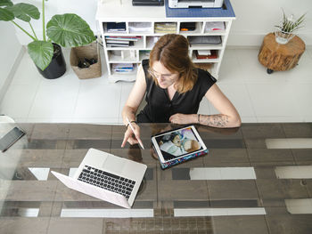 From above of female graphic designer using netbook and tablet with stylus while working on project at table in creative studio