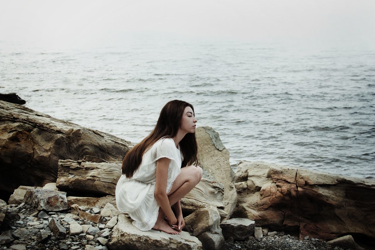 YOUNG WOMAN SITTING ON ROCK BY SEA