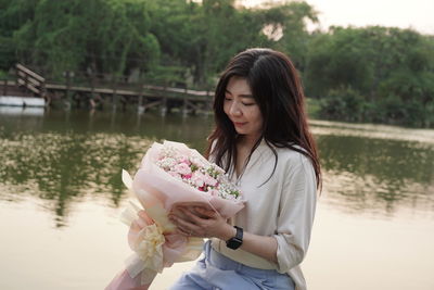 Young woman holding bouquet flowers while standing against the lake