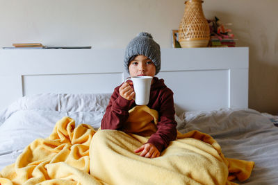 Girl wearing grey warm hat sits on the bed under warm blanket