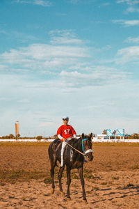 Rear view of young boy horse rider standing on field against clear sky