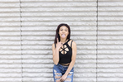 Portrait of young woman gesturing and sticking out tongue against wall