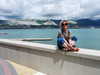 Side view of woman sitting on retaining wall by sea against sky