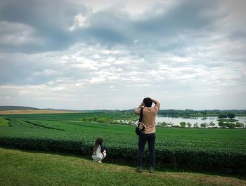 Rear view of mid adult man photographing while daughter crouching on grassy field against cloudy sky