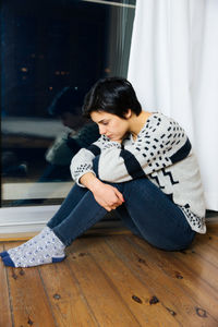 Full length of depressed woman sitting on hardwood floor by window at home