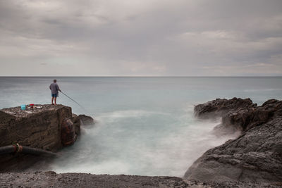 Man fishing at sea shore against cloudy sky