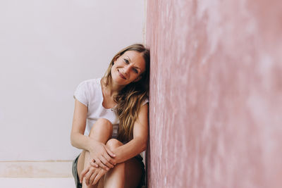 Blond, caucasian woman leaning against a colored wall and looking at the camera