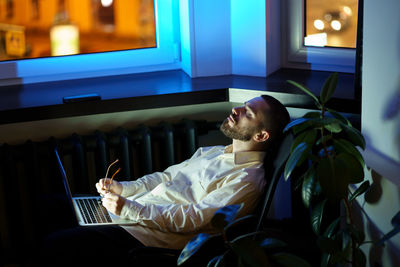 Exhausted young businessman or programmer sit with closed eyes in dark office with laptop overtime