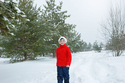 A teenager in a red sweater and hat walks in the winter  near the pines in the snow, smiling.