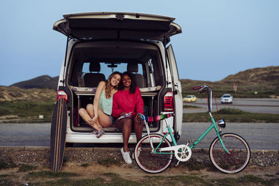 Cheerful women smiling at camera while sitting on trunk of van next to bike and skateboard in camping at dusk