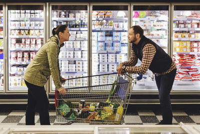 Side view of couple talking while leaning on shopping cart at refrigerated section