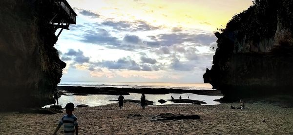 People on rocks by sea against sky during sunset