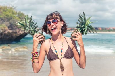 Happy young woman holding pineapples at beach against sky