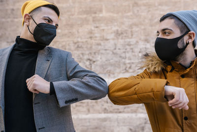 Male friends wearing protective face mask and knit hat giving elbow bump while standing against wall