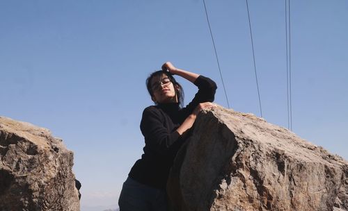 Low angle portrait of young woman standing on rock against clear sky
