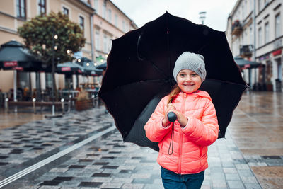 Little smiling happy girl holding big umbrella walking in a downtown on rainy gloomy autumn day