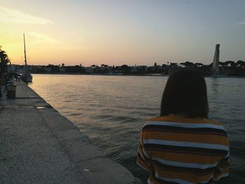 Rear view of woman standing by river against sky during sunset