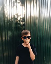 Portrait of boy eating ice cream cone while standing against corrugated iron