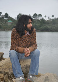 A young man with long hair and beard looking sideways while sitting on stone by the lake