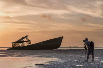 Side view of man photographing abandoned boat at beach against cloudy sky during sunset