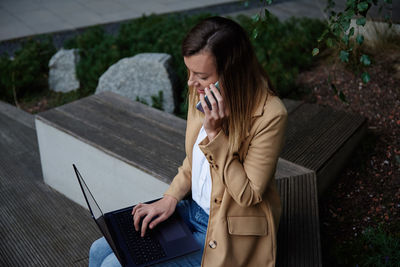 Woman sitting on bench in city street, using laptop and smartphone