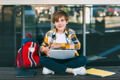 Portrait of boy using laptop while sitting on bench against modern building