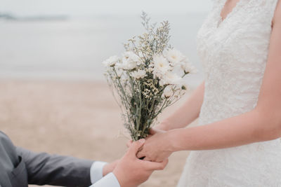 Midsection of groom holding hands of bride with bouquet at beach