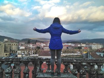 Rear view of woman with arms outstretched standing at balcony against cloudy sky