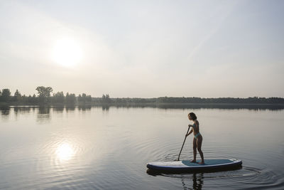 Woman doing standup paddleboarding in lake at sunset