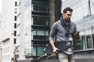 Businessman carrying skateboard, using smartphone and earphones