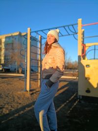 Side view of young woman looking away while standing against clear blue sky in playground during sunset