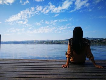 Rear view of mid adult woman sitting by lake against sky
