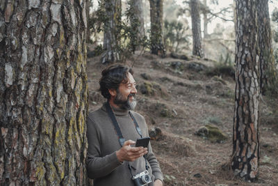 Smiling man holding phone standing by tree in forest
