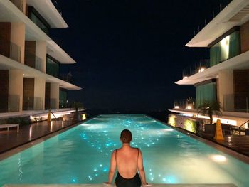 Rear view of woman sitting by illuminated swimming pool at night
