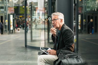 Senior male commuter talking through in-ear headphones while sitting by bags at railroad station