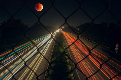 Light trails on chainlink fence against sky at night