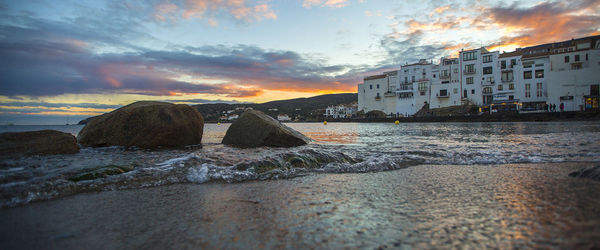 Panoramic view of rocks in sea by buildings against sky during sunset