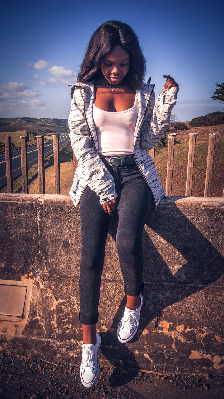 FULL LENGTH PORTRAIT OF A YOUNG WOMAN STANDING AGAINST RAILING