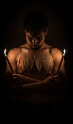 Shirtless man holding lit candle with wax on body against black background