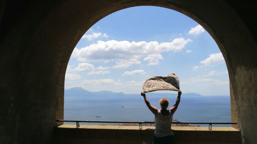 Rear view of woman holding scarf while standing in arch balcony against sky