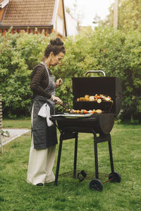 Full length side view of senior woman preparing meal on barbecue grill during dinner party at back yard