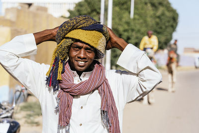 Positive arab male wearing traditional turban standing on street in small settlement on sunny day and looking at camera