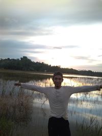 Portrait of man with arms outstretched standing against lake during sunset