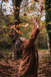 Medium shot of a brunette woman throwing dry leaves into the air in a forest in autumn.