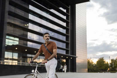 Contemplative businessman wheeling with bicycle in front of building