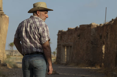 Rear view of adult man in cowboy hat and shirt against abandoned building