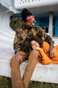 Happy black man sitting and woman lying down in stylish casual outfits cuddling on concrete bench